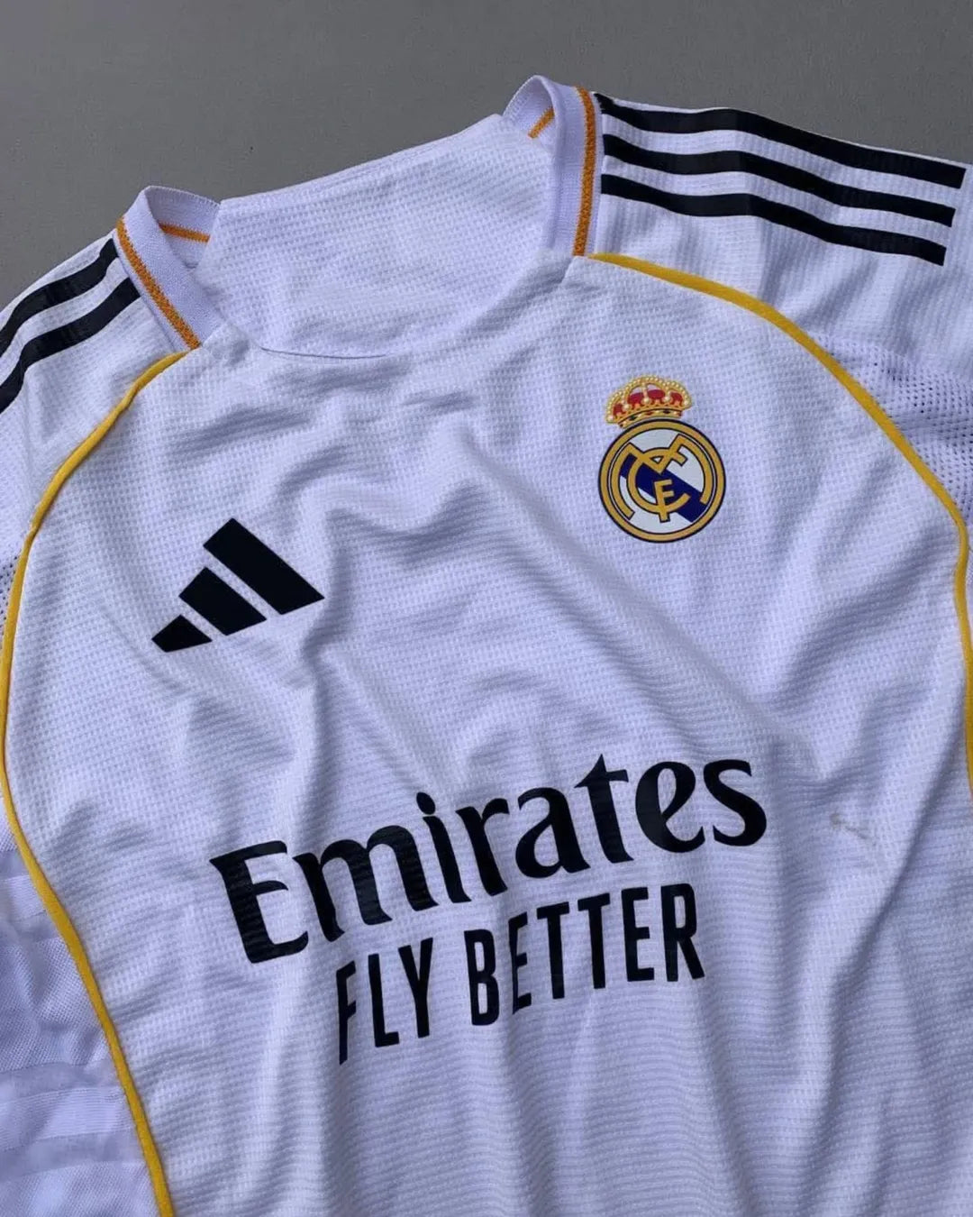 Real Madrid 2025/26 home jersey with classic white design, black adidas stripes, golden-yellow trim, Emirates Fly Better sponsor, and embroidered club crest – official fan and player kit